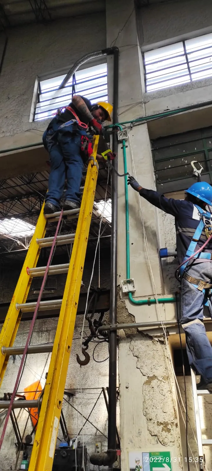 Dos técnicos con casco y arnés de seguridad instalan una tubería de conducción para cableado en el interior de una bodega industrial, uno en lo alto de una escalera y otro sosteniendo el tramo inferior