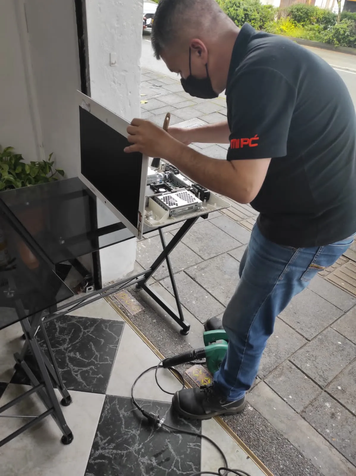 Técnico de MiPC Tecnología con tapabocas repara un computador todo en uno, con la parte trasera abierta y la tarjeta madre a la vista, sobre una mesa plegable en la calle