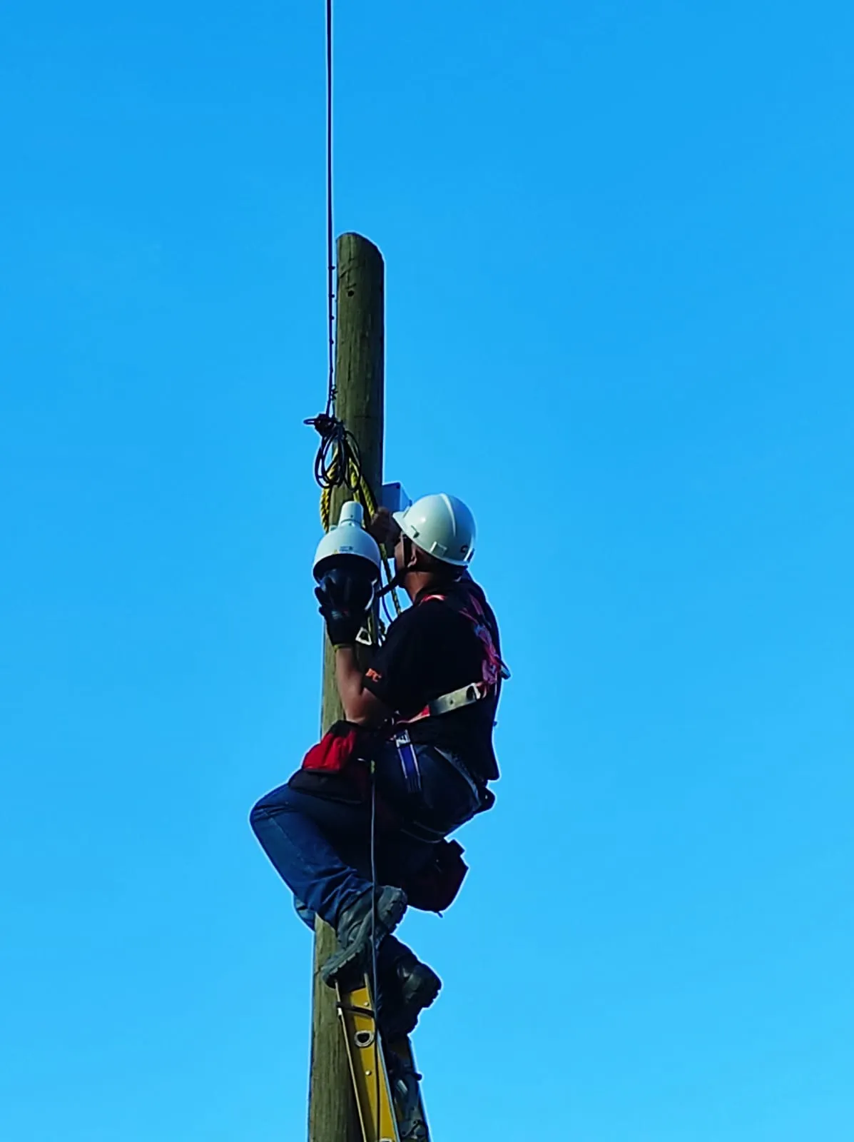 Técnico de MiPC Tecnología con casco blanco y arnés de seguridad instala una cámara domo de videovigilancia en lo alto de un poste de madera, contra un cielo despejado